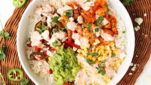 A bowl filled with rice, grilled shrimp, sautéed peppers, corn, guacamole, creamy sauce, and fresh cilantro, arranged in sections on a wicker placemat with sliced jalapeños and cilantro leaves around.