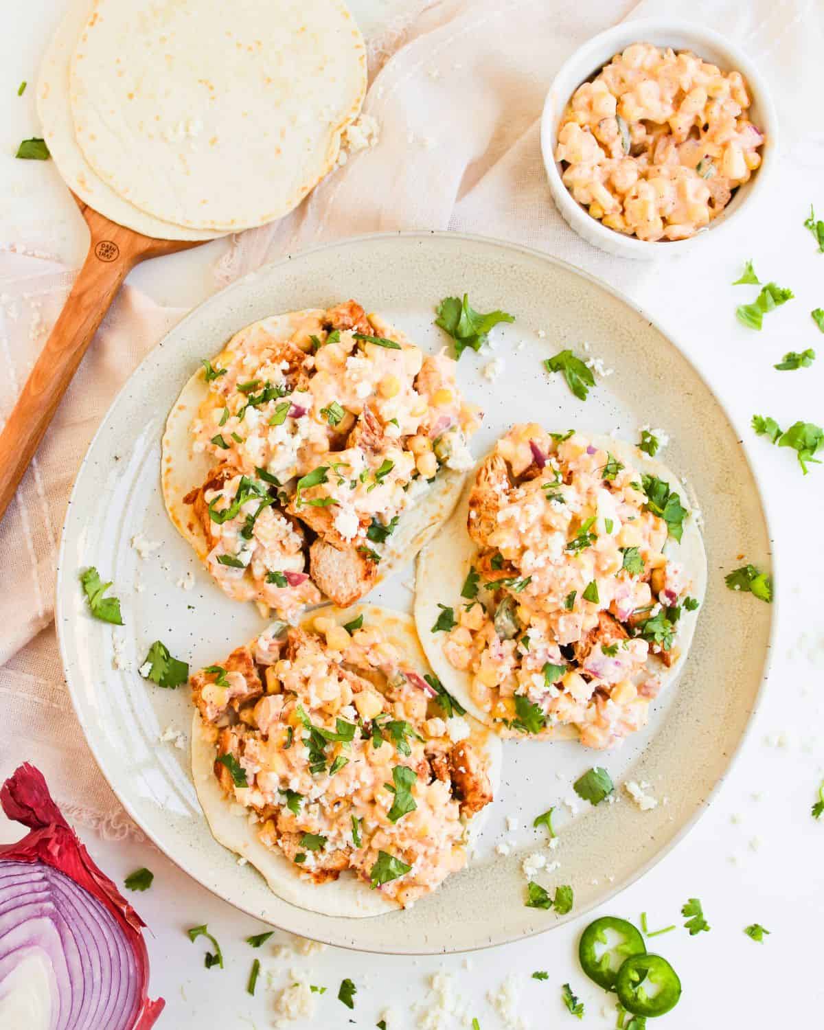 A plate with four tacos filled with a creamy corn mixture, chopped red onions, cheese, and fresh cilantro. A bowl of the corn mixture, sliced jalapeño, a stack of tortillas, and red onion are nearby.