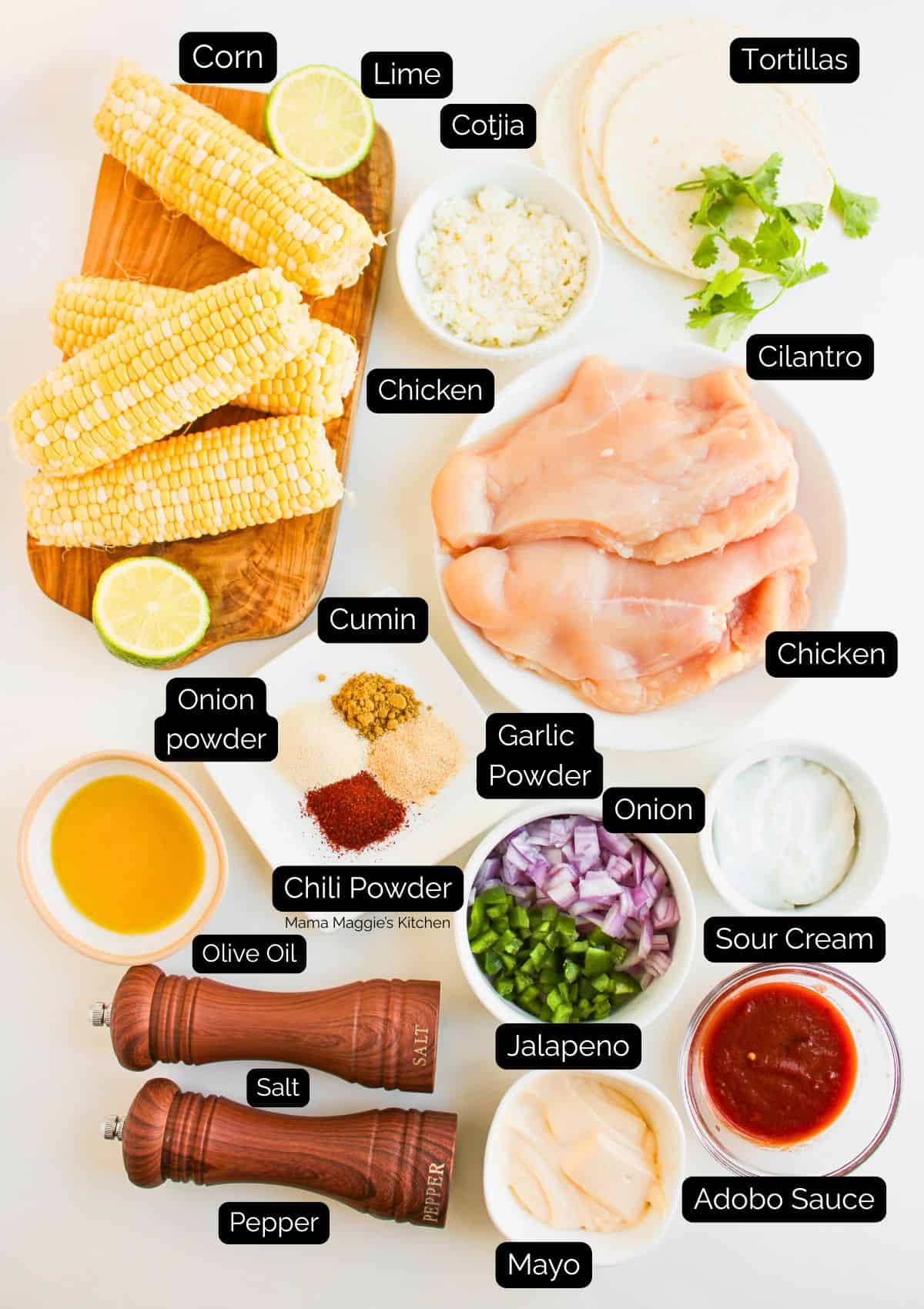 Top-down view of ingredients labeled for a Mexican dish: corn, lime, cotija cheese, tortillas, cilantro, raw chicken breasts, onion, jalapeño, sour cream, adobo sauce, mayo, olive oil, chili powder, spices, salt, and pepper.