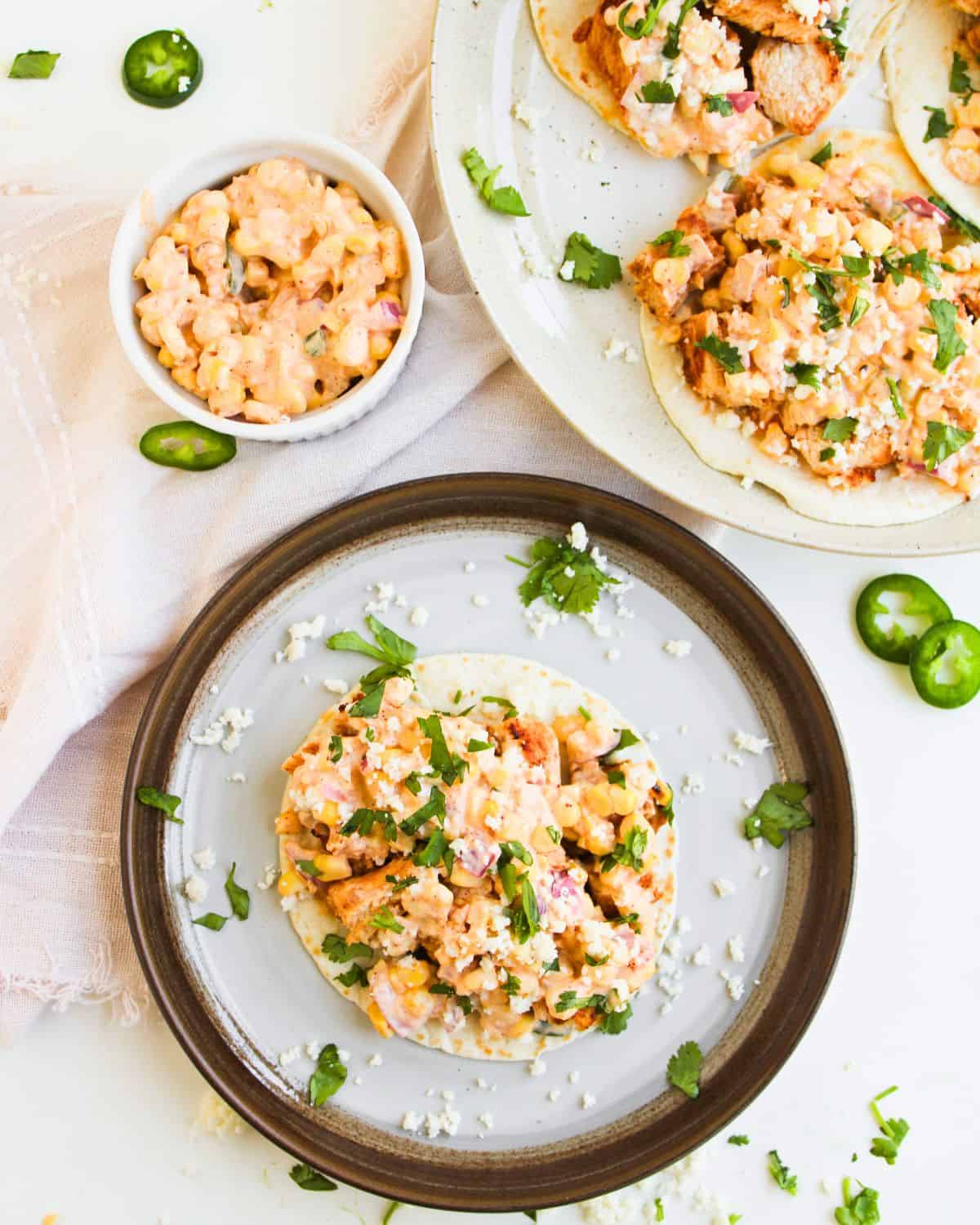 A plate with a tostada topped with creamy corn salsa, grilled chicken, crumbled cheese, and chopped cilantro. Nearby are more tostadas on another plate, a small bowl of corn salsa, jalapeño slices, and a light napkin.
