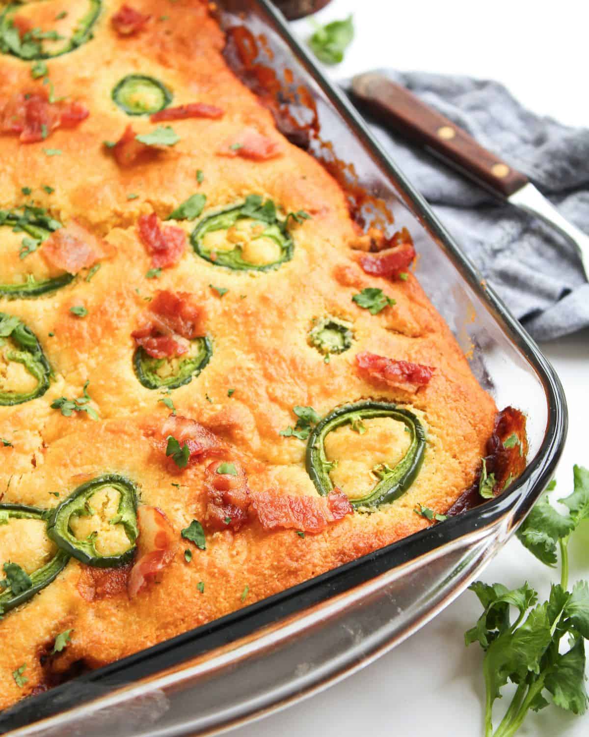 A baked cornbread casserole in a glass dish, topped with sliced jalapeños, bits of crispy bacon, and sprinkled herbs. A gray napkin, knife, and fresh cilantro are nearby on a white surface.