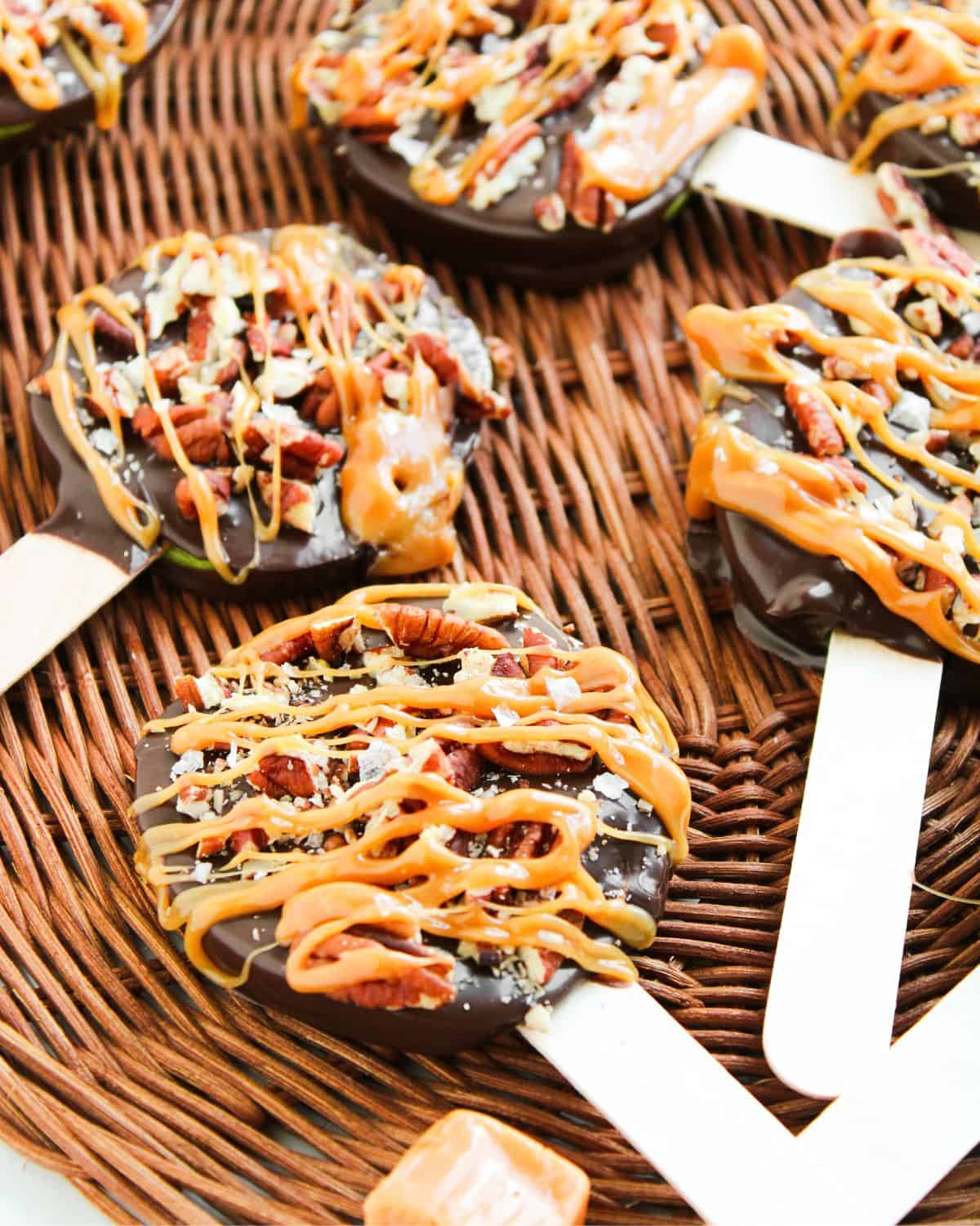 Chocolate-covered apple slices on popsicle sticks, topped with chopped pecans and drizzled with caramel, arranged on a wicker surface.