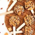 Five chocolate-covered caramel and pecan lollipops on wooden sticks, drizzled with caramel, are arranged on a woven placemat. Caramel candies and pecan pieces are scattered around them.