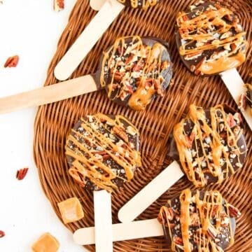 Five chocolate-covered caramel and pecan lollipops on wooden sticks, drizzled with caramel, are arranged on a woven placemat. Caramel candies and pecan pieces are scattered around them.