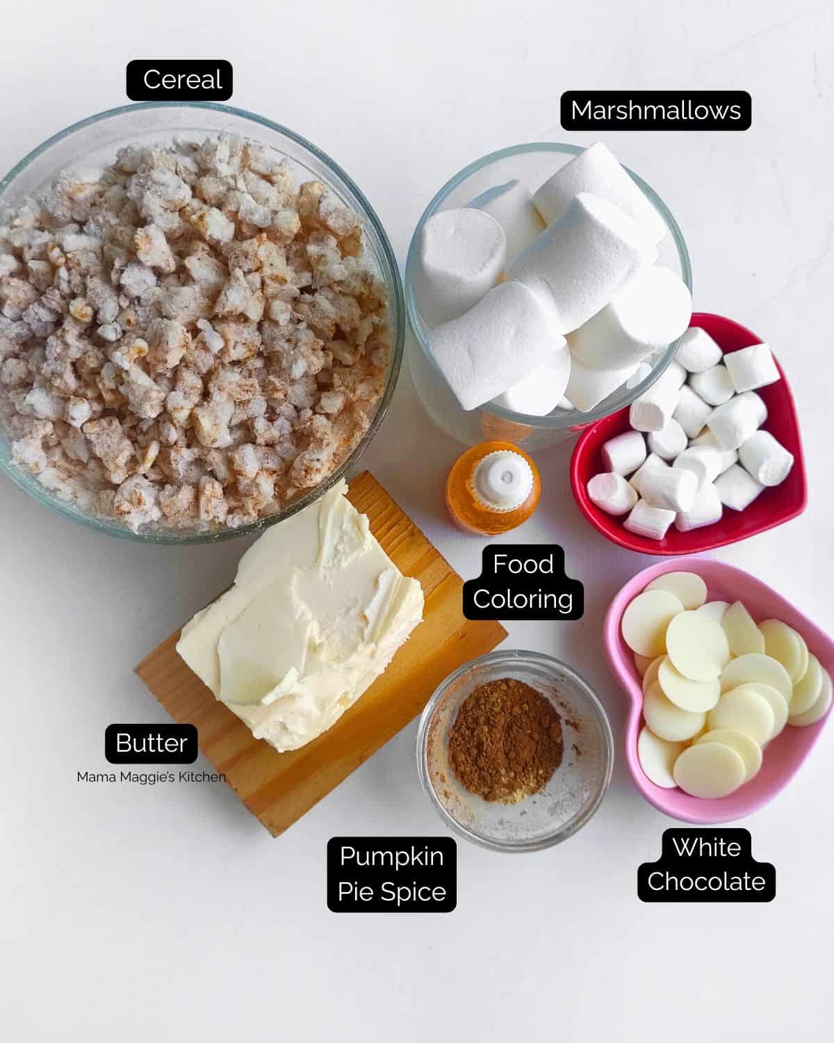 Bowls of cereal, large and small marshmallows, a dish of butter, a small bowl of pumpkin pie spice, white chocolate disks, and an orange food coloring bottle are arranged on a white surface. Labels identify each ingredient.