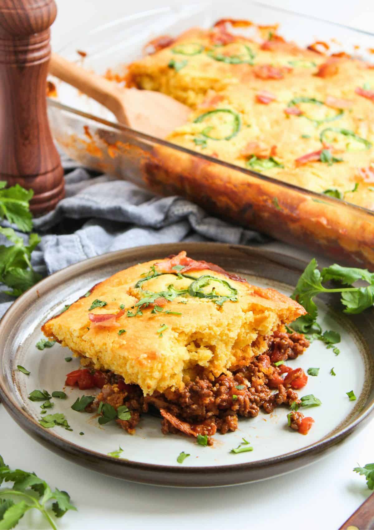 A plate with a serving of Mexican Cornbread Casserole With Jiffy, filled with seasoned ground meat and topped with sliced jalapeños and chopped cilantro. The casserole dish and a wooden serving spoon are visible in the background.
