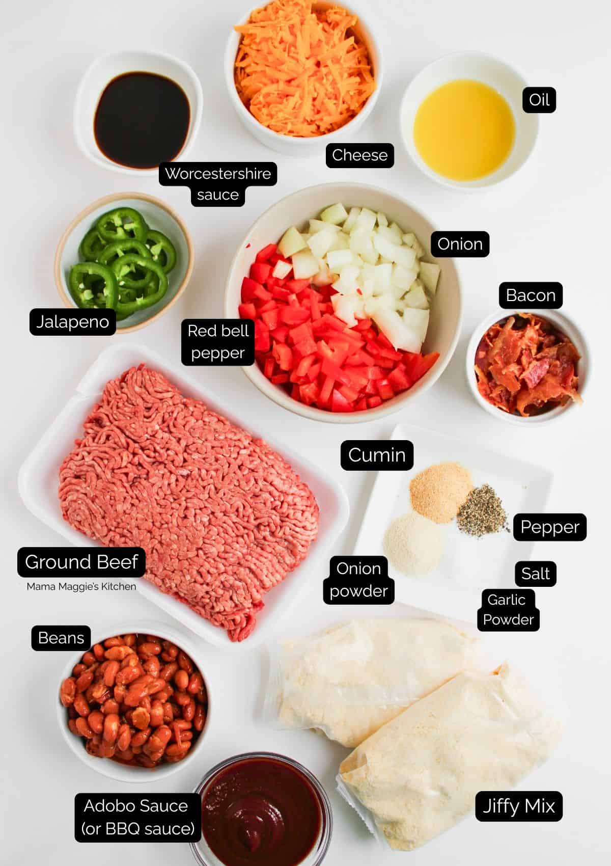Top-down view of ingredients for a casserole, including ground beef, onion, tomato, bell pepper, garlic powder, cumin, kidney beans, corn, Jiffy Mix, shredded cheese, Worcestershire sauce, and adobo sauce.