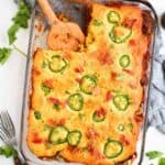 A glass dish filled with Mexican Cornbread Casserole With Jiffy, topped with jalapeno slices and bits of red pepper, with a wooden spatula resting in the casserole. Fresh cilantro and forks are placed nearby.