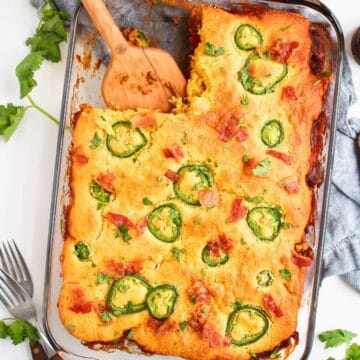 A glass dish filled with Mexican Cornbread Casserole With Jiffy, topped with jalapeno slices and bits of red pepper, with a wooden spatula resting in the casserole. Fresh cilantro and forks are placed nearby.