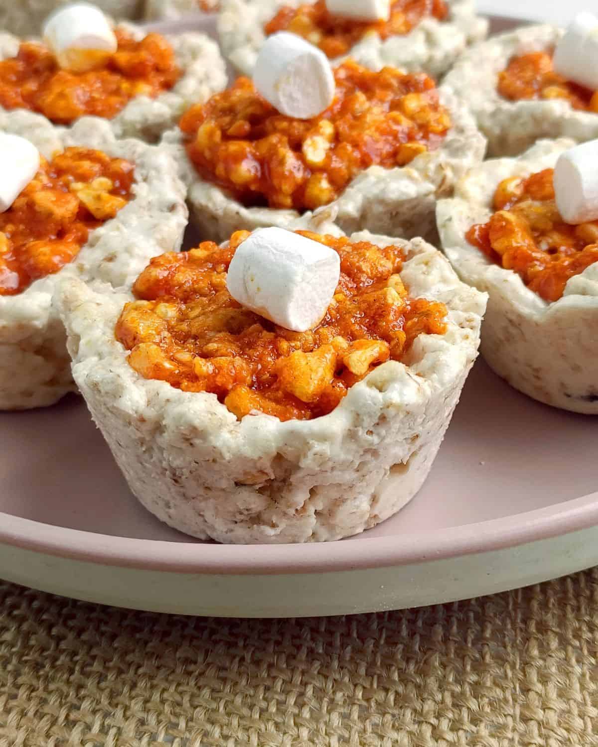 Mini dessert cups with a textured oat and nut base, filled with a sticky orange mixture, each topped with a small white marshmallow, arranged on a pink plate.