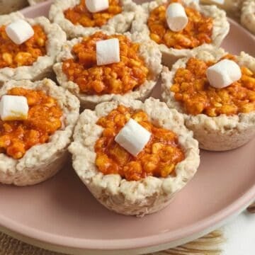 A plate of Pumpkin Pie Spice Rice Krispie Treats shaped like cups, filled with an orange mixture and topped with a mini marshmallow, arranged neatly on a light pink dish.