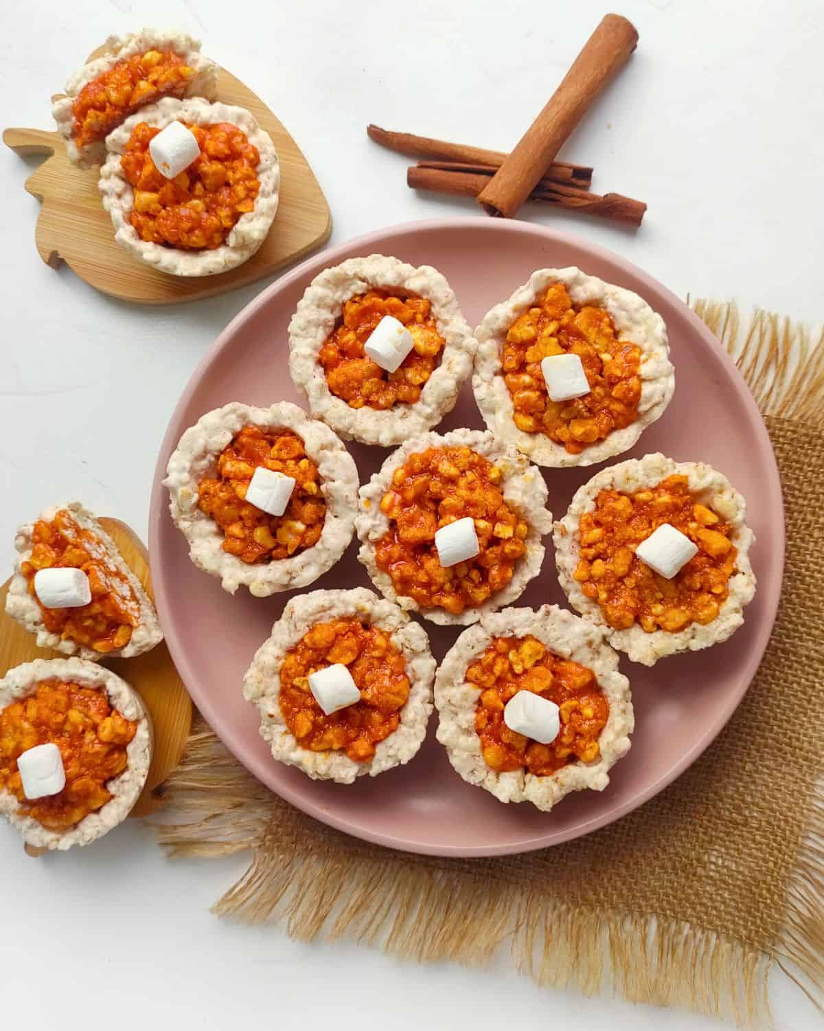 Pumpkin rice krispie treats with orange toppings, served on a pink plate, decorated with marshmallows, cinnamon sticks, and a woven cloth.