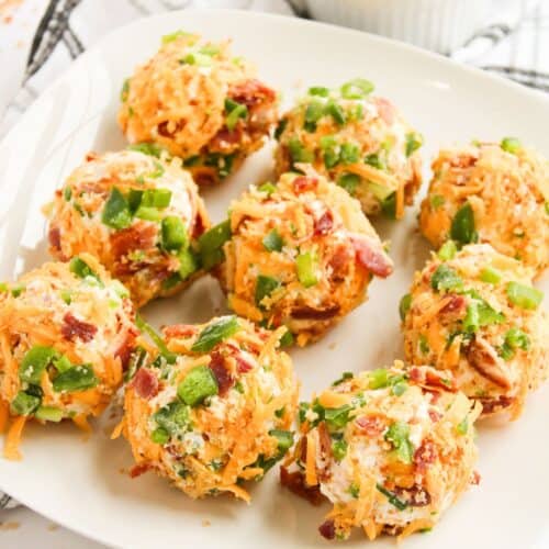A white plate holds eight Bacon Jalapeno Cheese Balls coated in shredded cheese, chopped green onions, and bits of bacon. A bowl of green onions sits in the background on a white cloth.