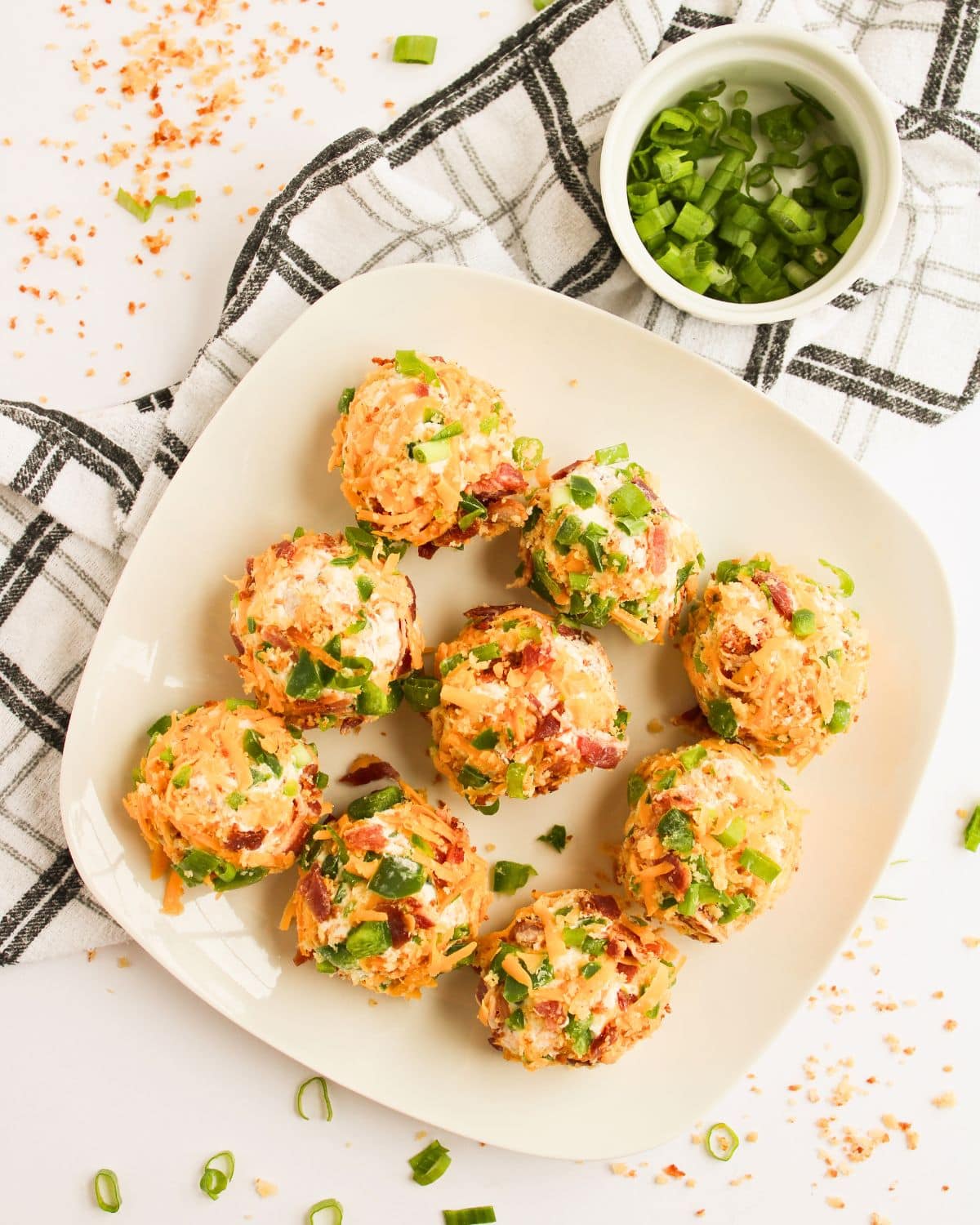 A plate with eight Bacon Jalapeno Cheese Balls garnished with chopped green onions and bacon bits. A small bowl of sliced green onions and a checkered cloth are nearby, with crumbs and green onions scattered on the white surface.
