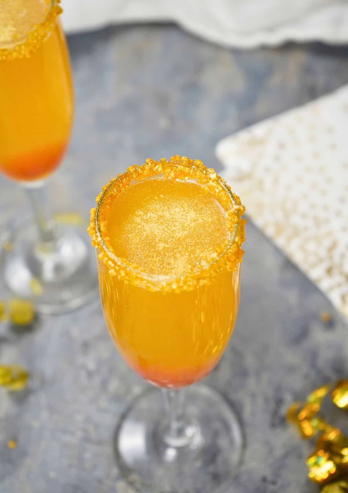 A Golden Champagne Cocktail fills a champagne flute, its orange-yellow sparkle and sugared rim glowing, while a hint of red rests at the bottom. The drink sits on a gray surface beside a gold-patterned napkin.