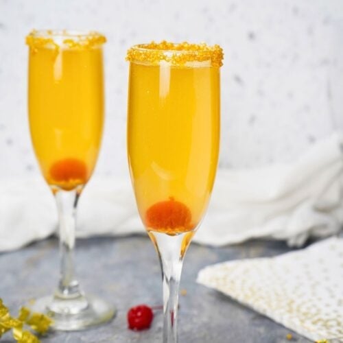 Two champagne flutes filled with a Golden Champagne Cocktail, each garnished with an orange sugar rim and a maraschino cherry. Decorative gold elements and a cherry are placed on the table nearby.