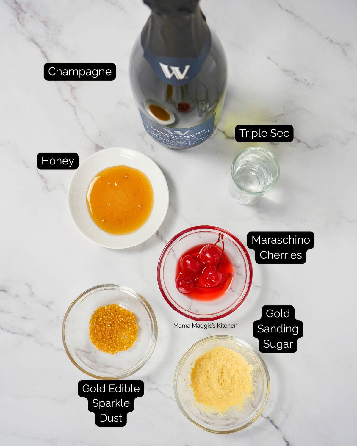Overhead view of ingredients on a white surface: a bottle of champagne, a glass of triple sec, a bowl of honey, maraschino cherries in a red bowl, gold edible sparkle dust, and gold sanding sugar. Each item is labeled.