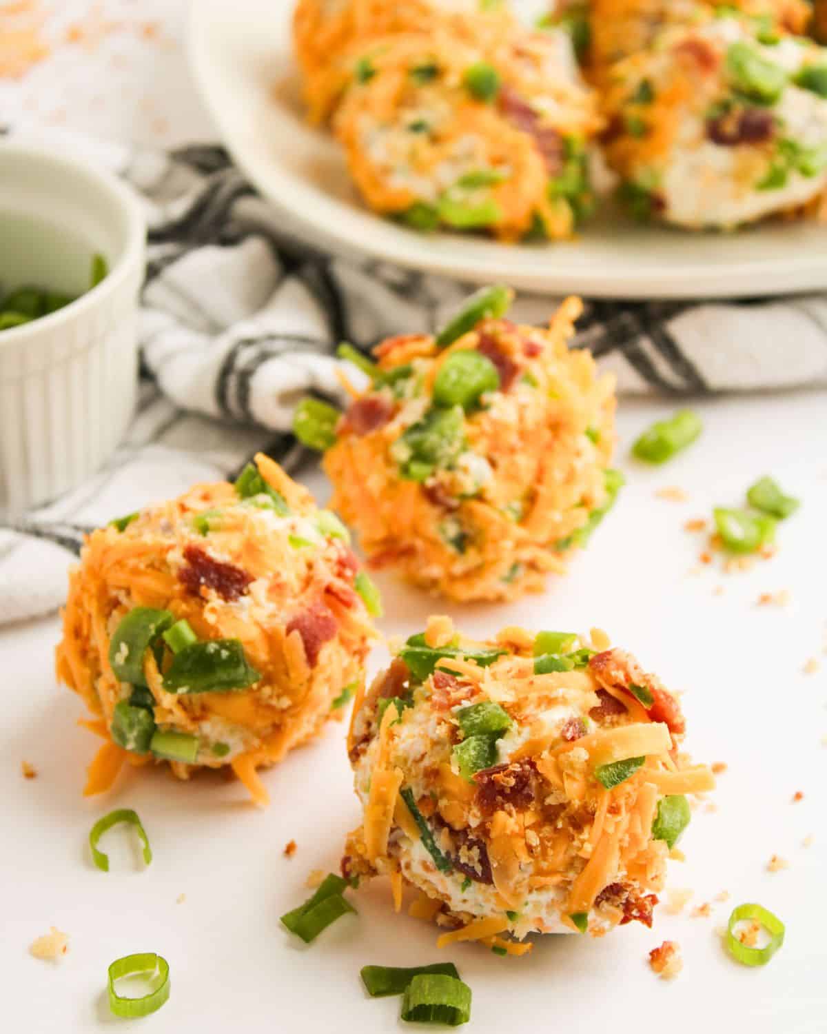 Three Bacon Jalapeno Cheese Balls coated with shredded cheese, chopped green onions, and bacon bits are arranged on a white surface, with a plate of more cheese balls and a small bowl of green onions in the background.