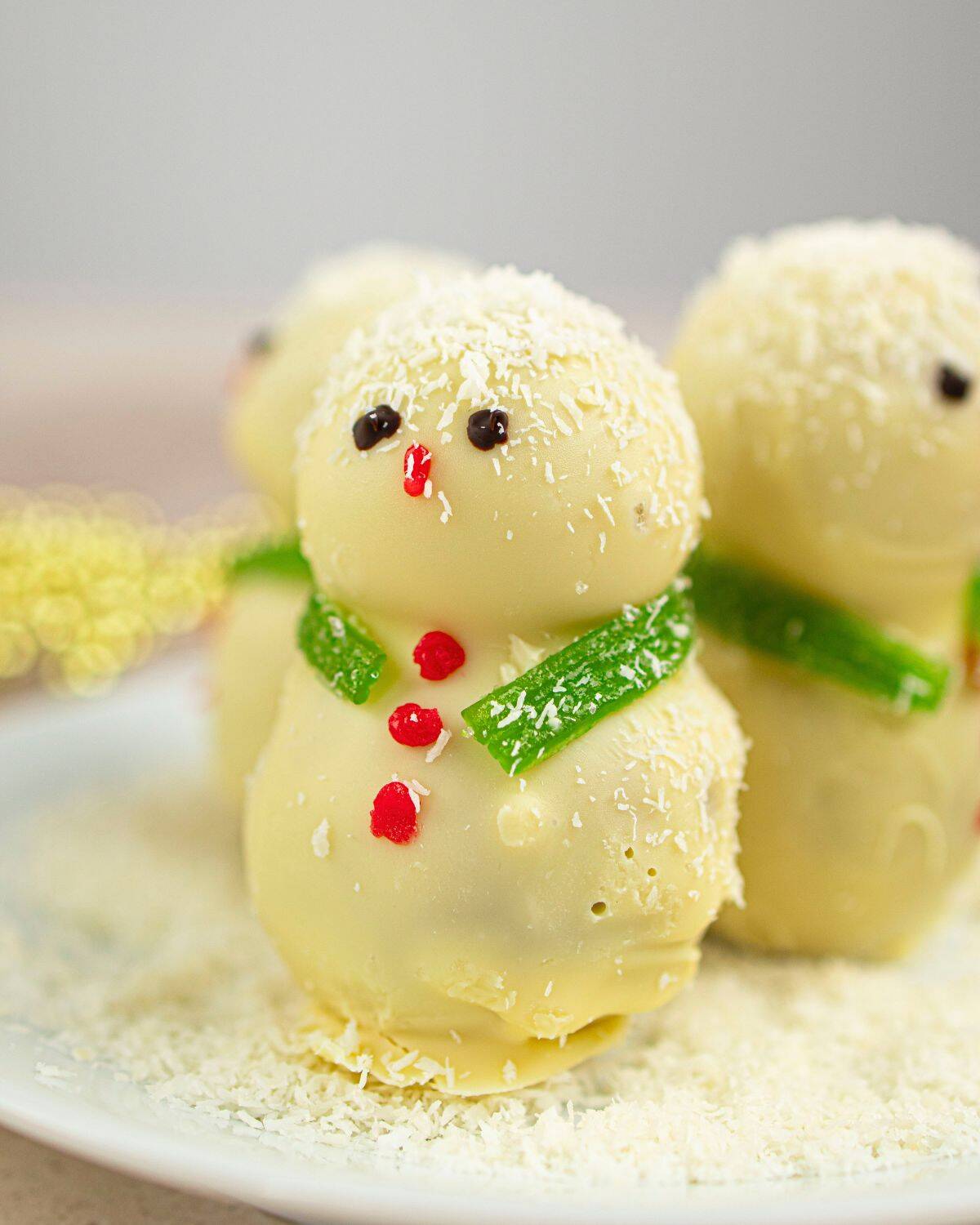 Three white chocolate snowmen decorated with red and green candy and chocolate details stand on a white plate sprinkled with shredded coconut, creating a festive, wintry appearance.