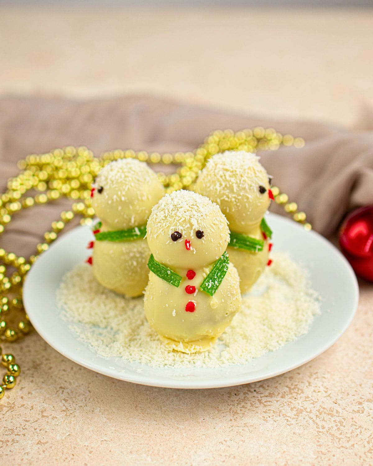Three snowman truffles decorated with green candy scarves and red buttons are arranged on a plate dusted with shredded coconut, with gold beads in the background.