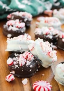 Peppermint Oreos dipped in chocolate and white chocolate are topped with crushed peppermint candies and arranged on a wooden surface, with whole peppermint candies nearby.