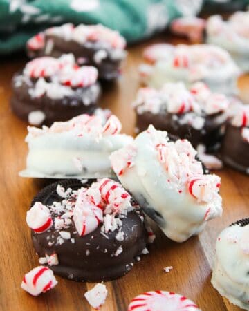 Peppermint Oreos dipped in chocolate and white chocolate are topped with crushed peppermint candies and arranged on a wooden surface, with whole peppermint candies nearby.