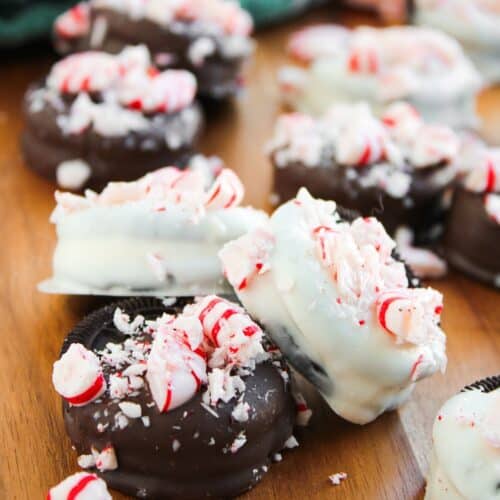 Peppermint Oreos dipped in chocolate and white chocolate are topped with crushed peppermint candies and arranged on a wooden surface, with whole peppermint candies nearby.