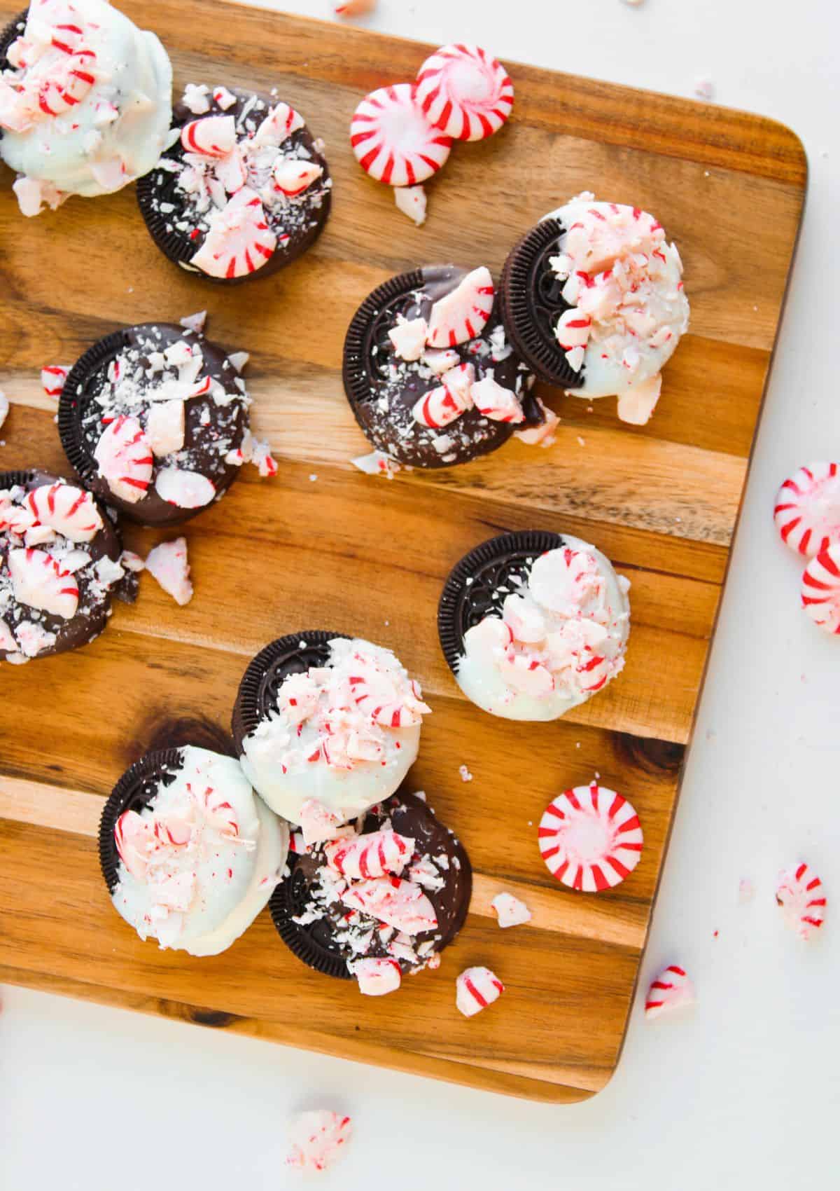 Peppermint Oreos topped with white frosting and crushed peppermint candies are arranged on a wooden board, surrounded by whole and broken peppermint candies.