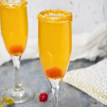 Two champagne flutes filled with a bright orange Golden Champagne Cocktail, each garnished with a sugar-rimmed edge and a red cherry at the bottom, set on a gray surface with gold and white decorations.
