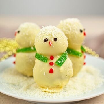 Three white chocolate Snowman Truffles with candy eyes, red candy buttons, green candy scarves, and coconut flakes sit on a white plate, with a soft, festive background.