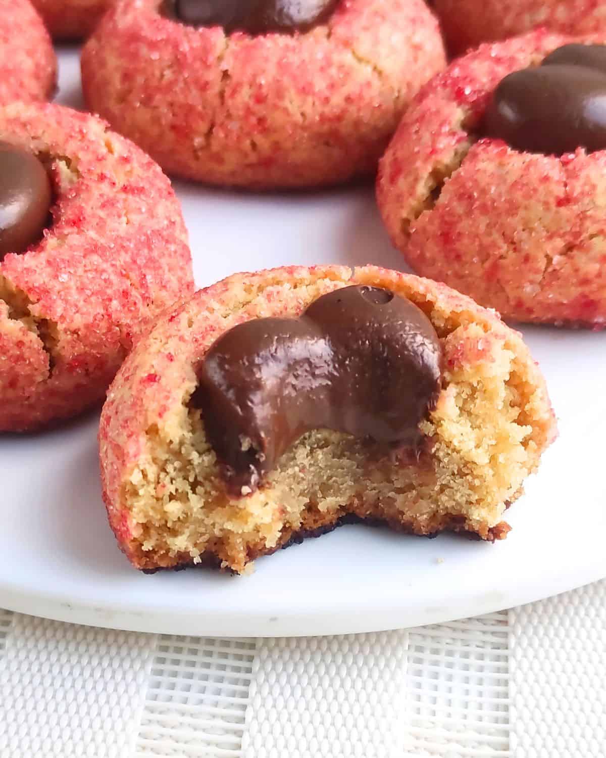 A close-up of a cookie with a chocolate center and a bite taken out, showing a soft, crumbly texture. Other similar cookies with pink sugar coating and chocolate centers are in the background on a white plate.