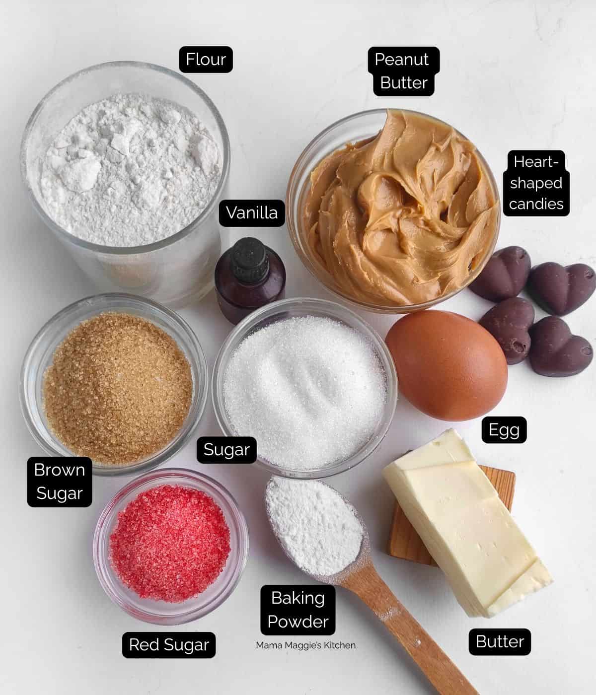 A flat lay of baking ingredients labeled: flour, peanut butter, heart-shaped candies, egg, butter, baking powder, red sugar, white sugar, brown sugar, and vanilla extract. All are arranged on a white surface.