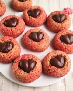 A plate of round cookies coated in pink sugar, each topped with a heart-shaped piece of chocolate in the center, arranged neatly on a white plate.