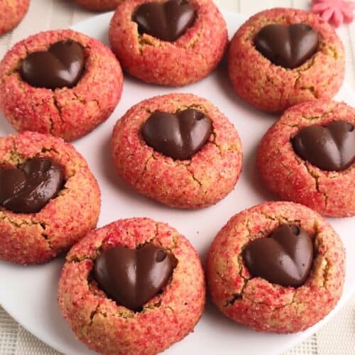 A plate of round cookies coated in pink sugar, each topped with a heart-shaped piece of chocolate in the center, arranged neatly on a white plate.