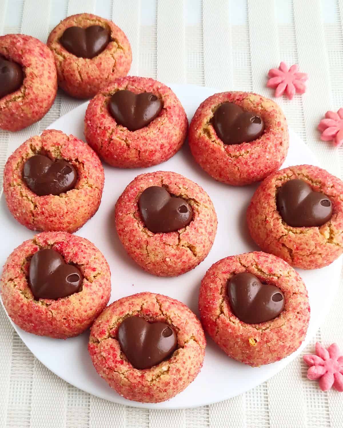 A white plate with ten pink Valentine's Day Peanut Butter Blossoms, each topped with a chocolate heart. The plate rests on a light-colored, textured surface, with pink flower decorations nearby.
