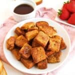 A white plate filled with golden brown Air Fryer French Toast Bites sits on a pink cloth, next to a bowl of syrup, a plate of fresh strawberries, and a slice of bread in the background.