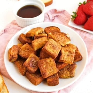 A white plate filled with golden brown Air Fryer French Toast Bites sits on a pink cloth, next to a bowl of syrup, a plate of fresh strawberries, and a slice of bread in the background.