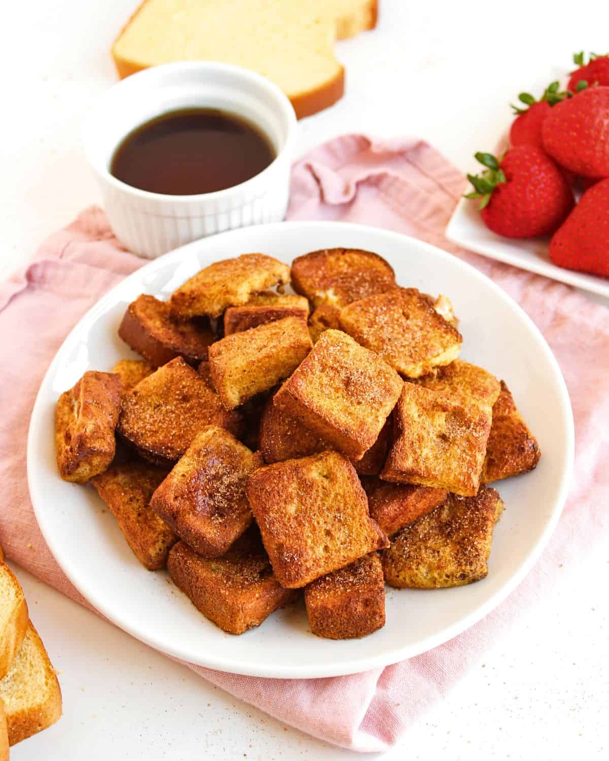 A white plate filled with golden brown Air Fryer French Toast Bites sits on a pink cloth, next to a bowl of syrup, a plate of fresh strawberries, and a slice of bread in the background.