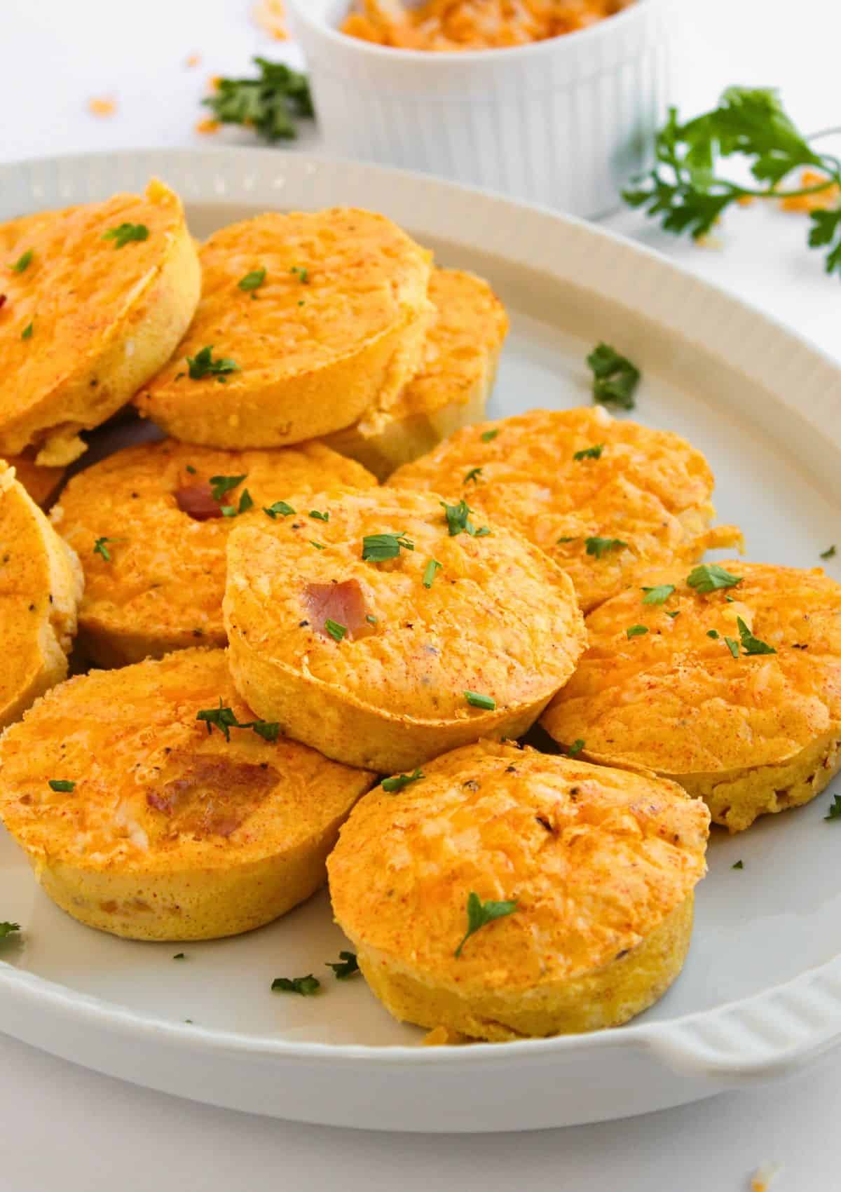 A white plate filled with several mini Ham and Cheese Egg Bites, golden brown and sprinkled with chopped parsley. Some muffins show bits of ham or cheese inside. A small ramekin and more parsley are seen in the background.