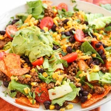 A plate of Dorito Beef Taco Salad topped with ground beef, lettuce, cherry tomatoes, black beans, corn, shredded cheese, tortilla chips, and a scoop of guacamole, served on a white dish with an orange napkin underneath.