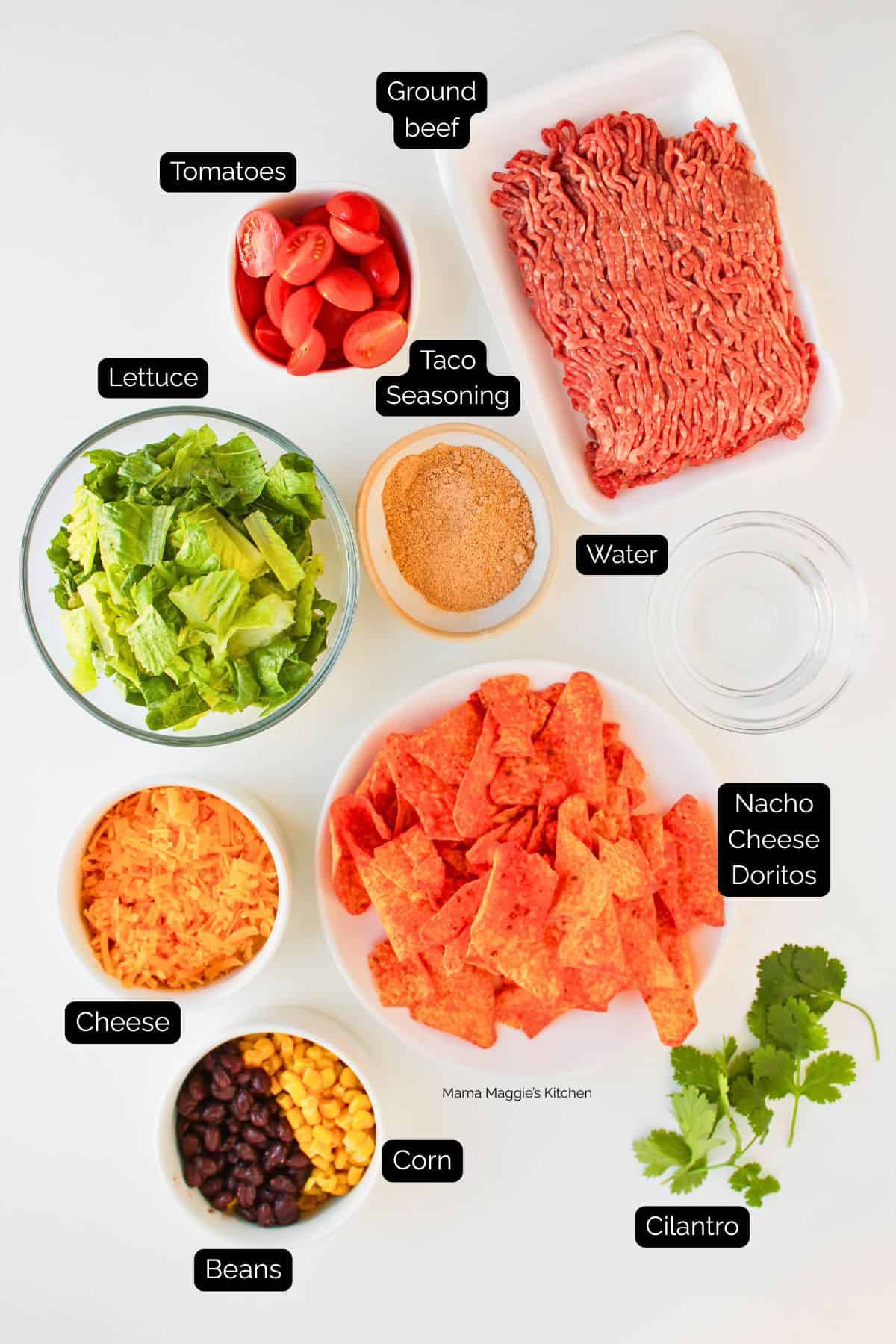 Flat lay of labeled Dorito Beef Taco Salad ingredients: ground beef, grape tomatoes, chopped lettuce, taco seasoning, water, shredded cheese, corn, black beans, nacho cheese Doritos, and cilantro on a white surface.