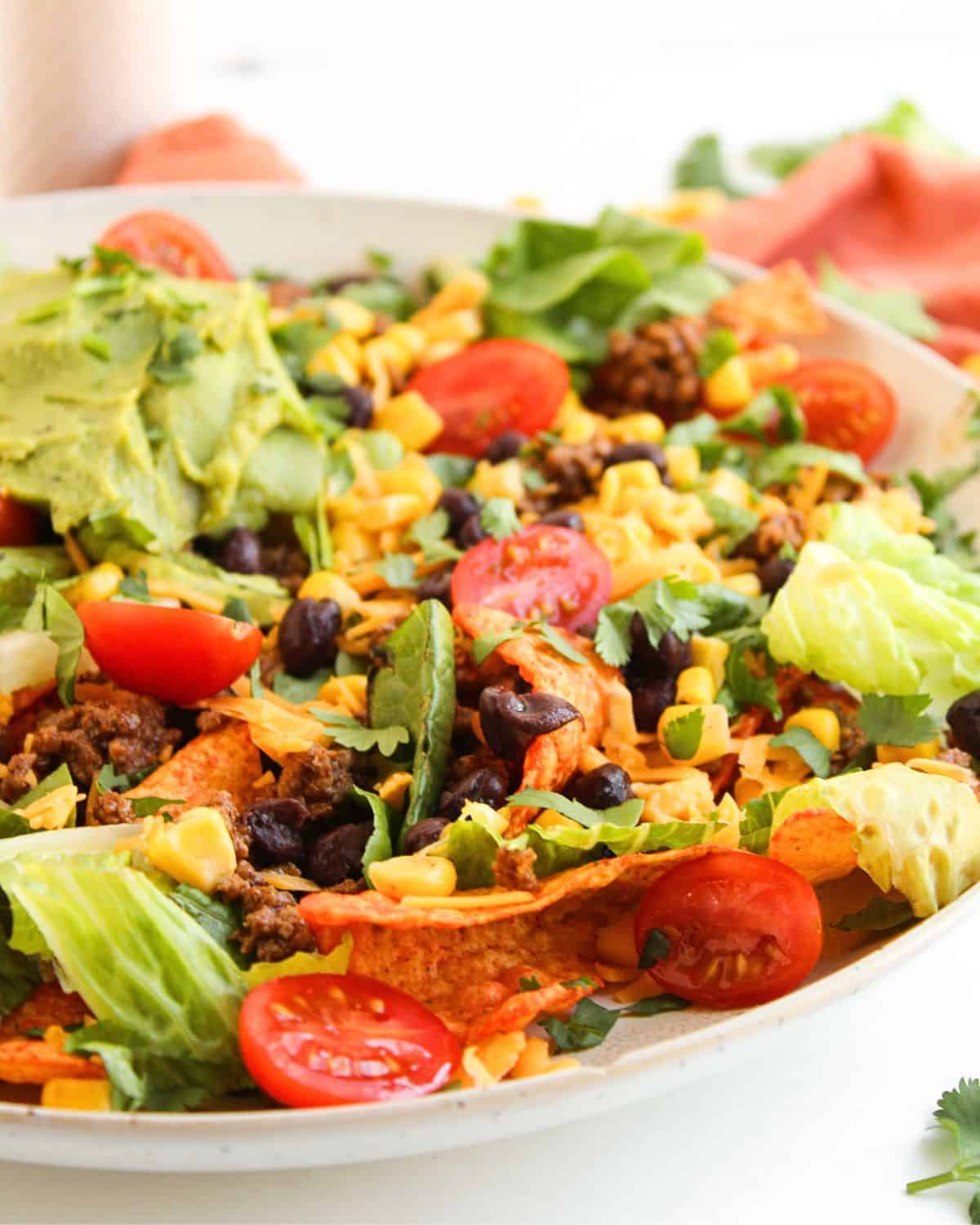 A plate of Dorito Beef Taco Salad with lettuce, cherry tomatoes, black beans, corn, seasoned ground beef, shredded cheese, tortilla chips, fresh cilantro, and a scoop of guacamole on top.