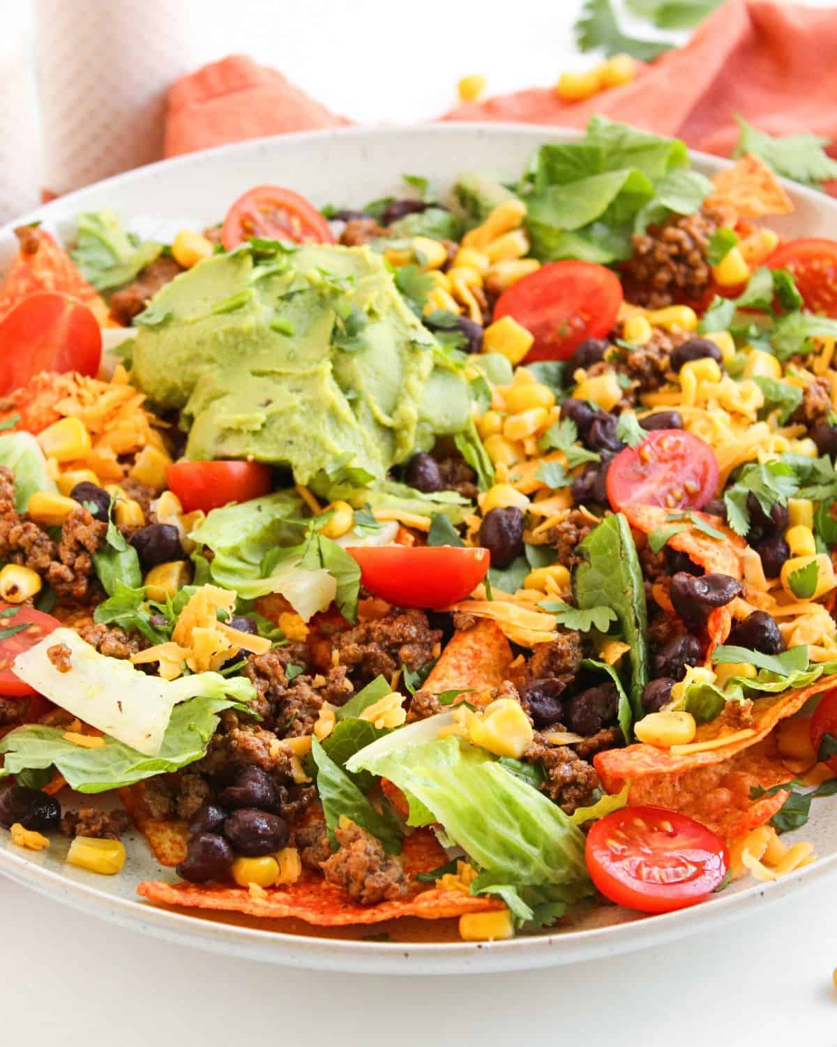 A flavorful Dorito Beef Taco Salad, this plate of loaded nachos is topped with seasoned ground beef, black beans, corn, shredded lettuce, cherry tomato halves, shredded cheese, and a generous scoop of guacamole in the center.