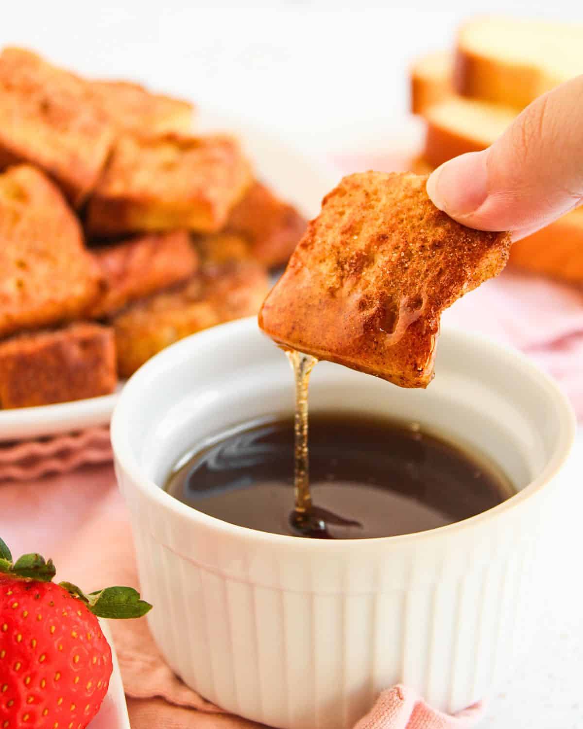 A hand dips a piece of golden-brown Air Fryer French Toast Bite into a small white ramekin filled with syrup. In the background, more French toast bites and a fresh strawberry are visible.