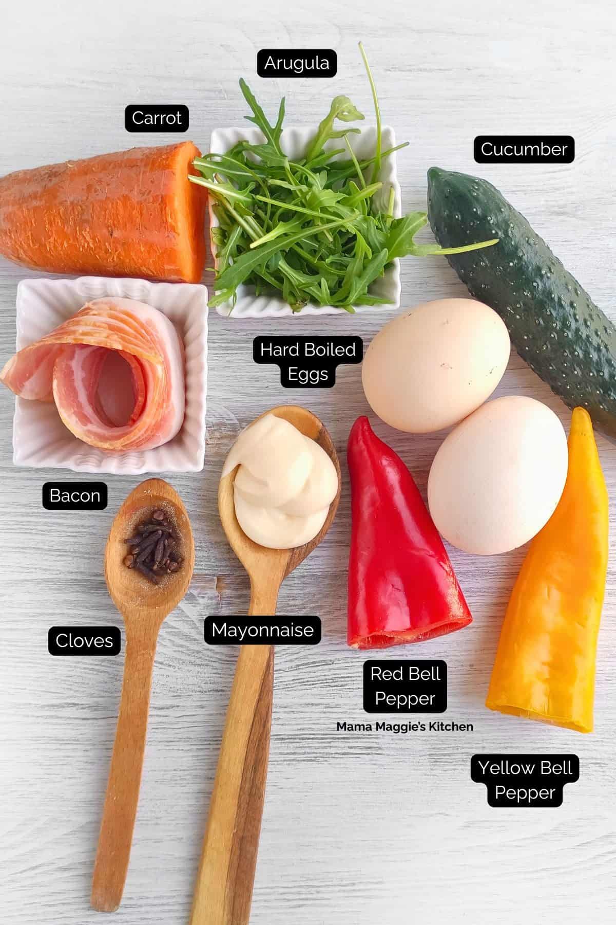 Labeled ingredients on a white surface: carrot, arugula, cucumber, hard-boiled eggs, bacon, mayonnaise, cloves, a red bell pepper, and a yellow bell pepper. Each item is arranged clearly with labels.