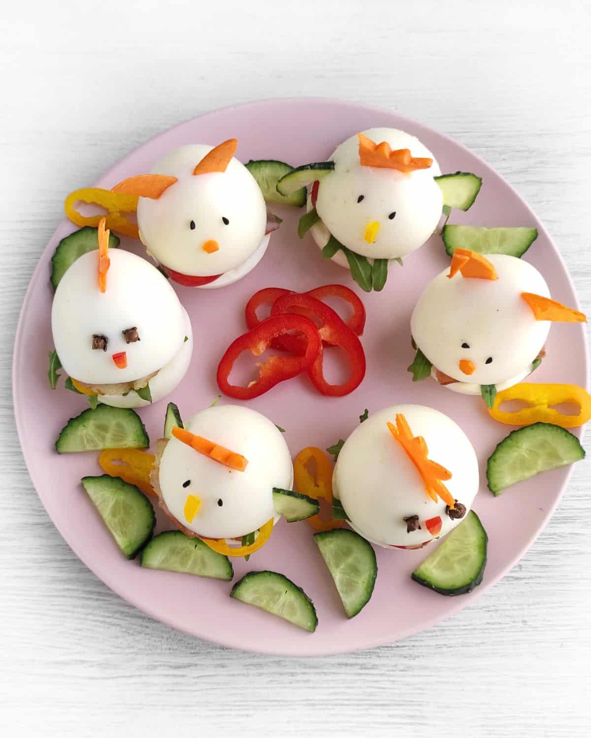 A pink plate features five Easter baby chick hard-boiled eggs, decorated with carrot pieces and cloves, surrounded by cucumber slices and bell pepper rings—perfect for a festive Easter sandwich platter.