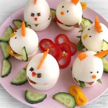 A pink plate with Easter sandwich baby chick and bunny hard-boiled eggs, decorated with carrot pieces and black pepper, surrounded by cucumber slices and bell pepper rings.