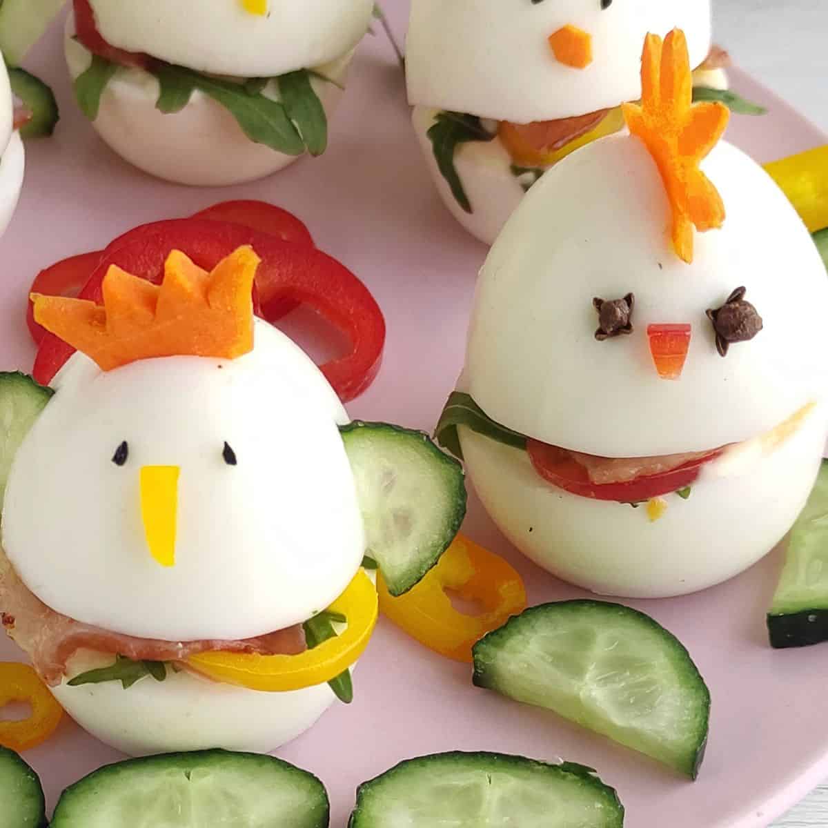 Two hard-boiled eggs decorated as Easter baby chicks with carrot combs, cheese beaks, and clove eyes. They sit on a plate beside cucumber slices and colorful bell peppers, perfect as a festive sandwich garnish.