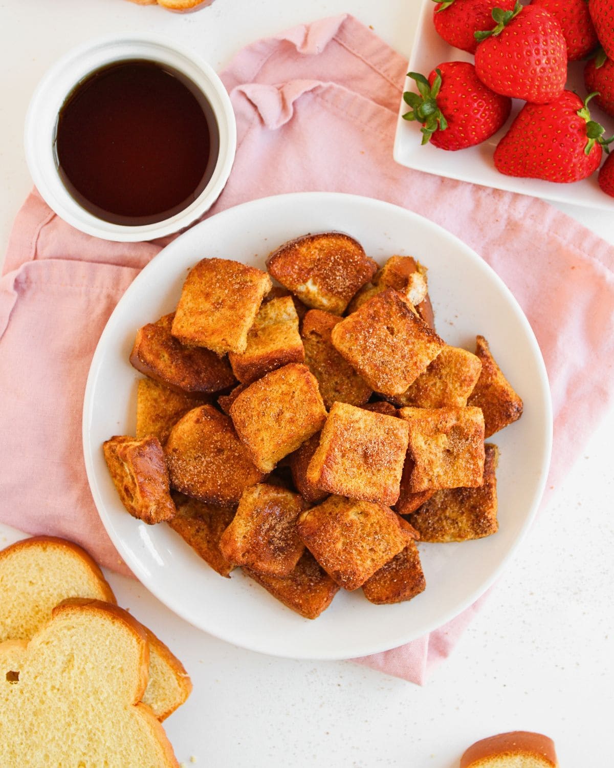 A white plate filled with Air Fryer French Toast Bites dusted in cinnamon sugar sits on a pink napkin, surrounded by fresh strawberries, syrup in a small bowl, and slices of bread on a white surface.