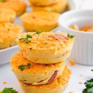 Three Ham and Cheese Egg Bites stacked on each other, garnished with parsley. More egg muffins are visible in the background on a plate, along with scattered cheese and parsley on a white surface.